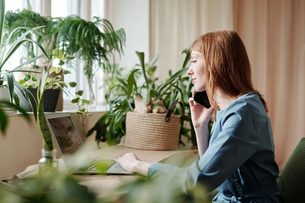 A woman on a phone call working at a laptop on a wooden table surrounded by potted plants