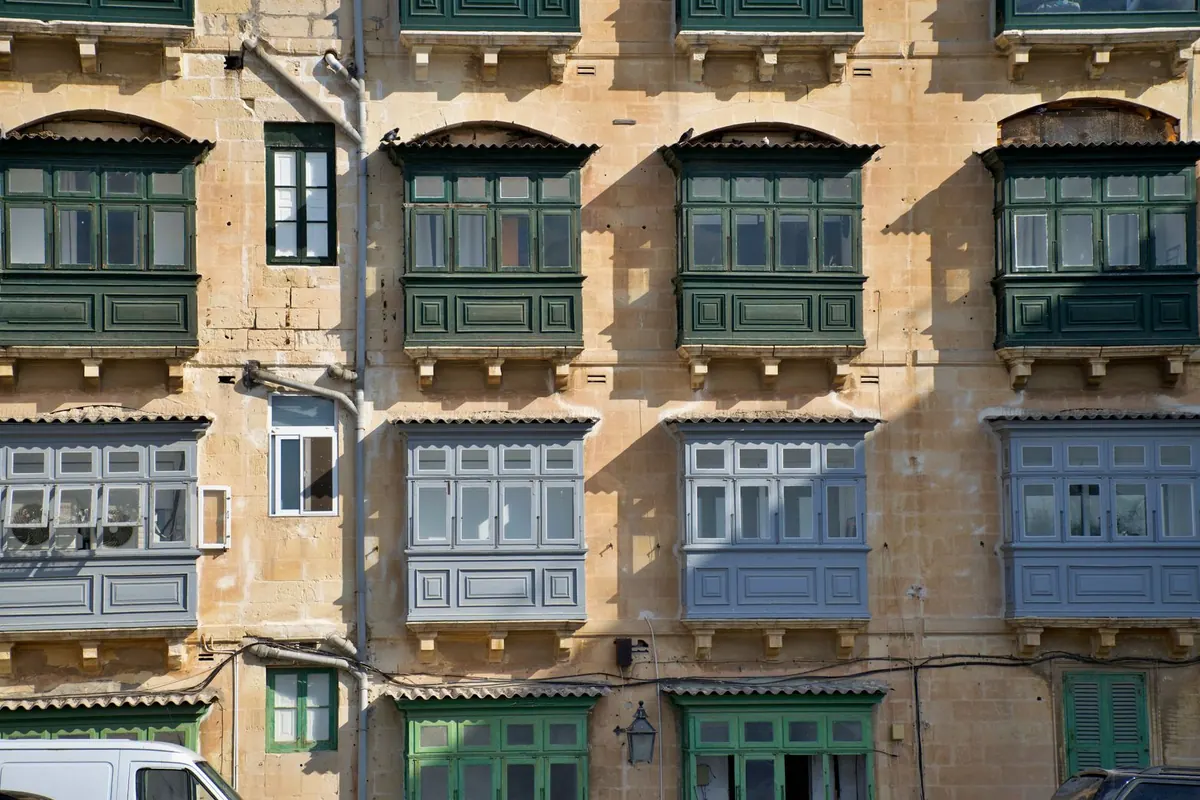 A sunlit Valletta limestone façade with rows of green and blue enclosed wooden balconies