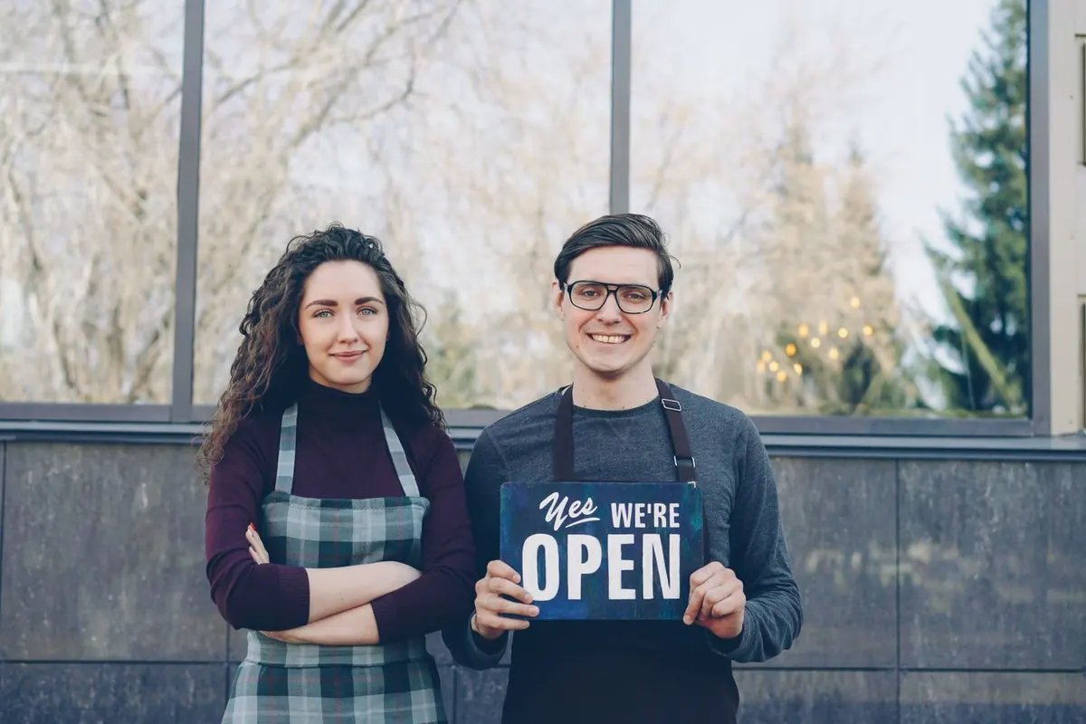 Two smiling small-business owners in aprons standing outside their shop holding a "Yes, we’re open" sign
