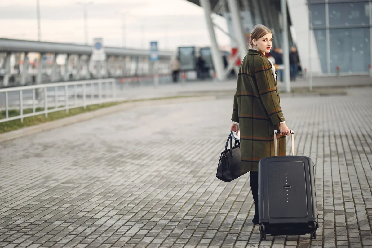 A woman in a long checked coat walking with a suitcase and handbag outside a modern airport terminal